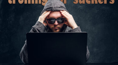 Pensive hacker in hoodie and sunglasses sits behind laptop in room against a dark wall